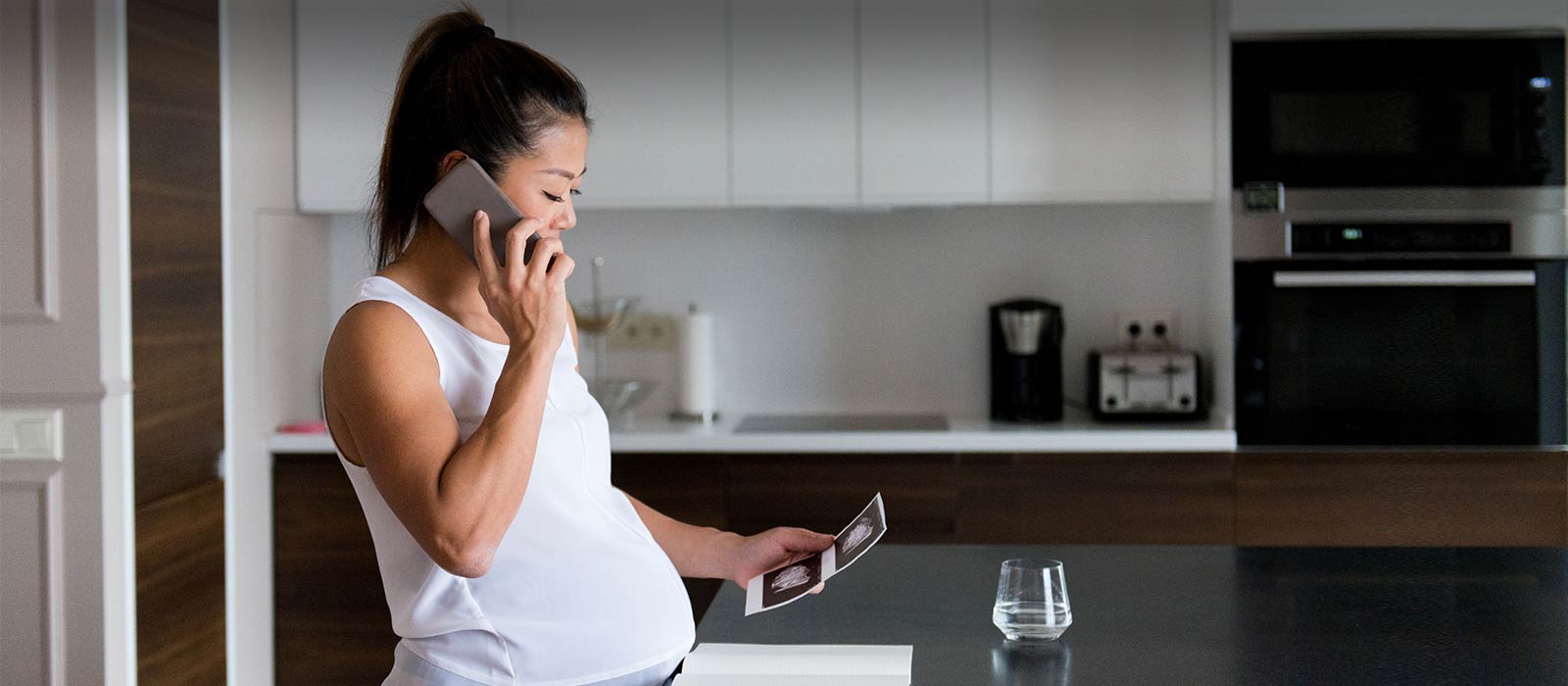 Pregnant woman on phone viewing sonogram image in kitchen.